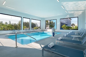 Una piscina interior moderna con paredes de vidrio transparente, zonas de descanso iluminadas por el sol y vistas a zonas verdes exteriores.