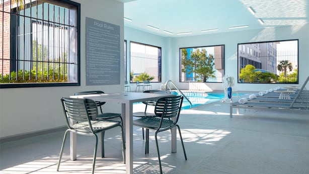 Un área de piscina interior moderna con una mesa y sillas, ventanas amplias y un área de descanso junto a la piscina con tumbonas.