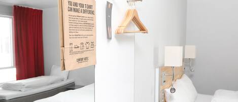 Habitación de hotel con dos camas, una cortina roja, un perchero de madera y un cartel sobre reciclaje de camisetas.