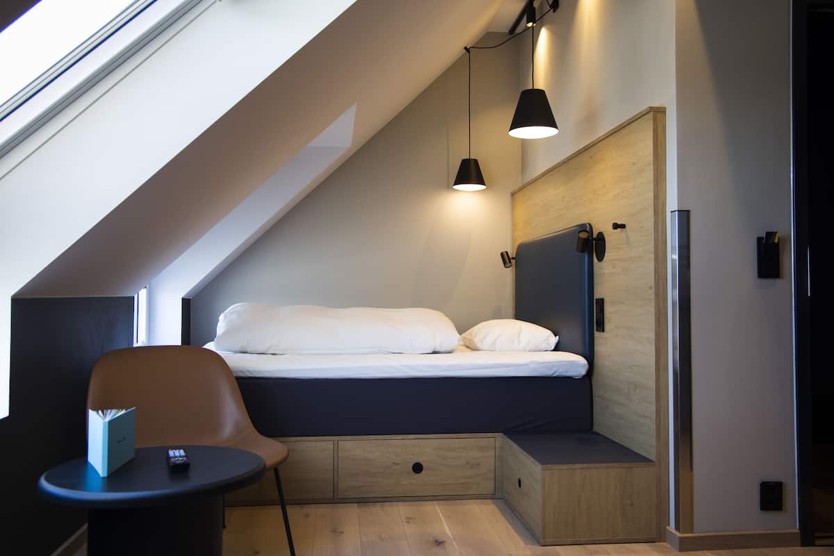 Une chambre à coucher de taille réduite, comprenant un lit, une chaise, une petite table et un banc intégré doté de tiroirs de rangement.