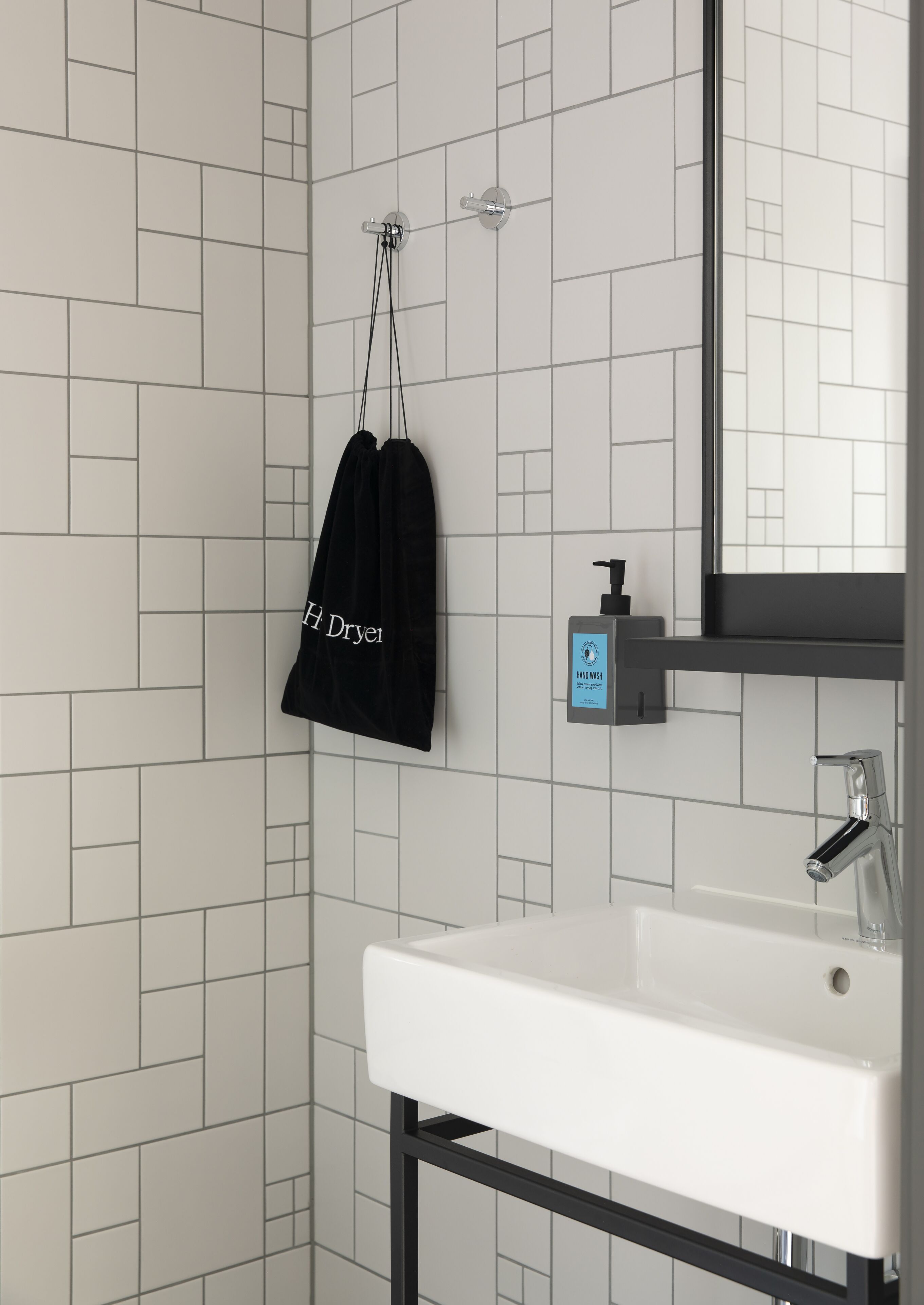 Ein Badezimmer mit einem weißen Waschbecken, einem Spiegel, einem Handseifen-Spender und einer schwarzen Tasche, die an der Wand hängt.