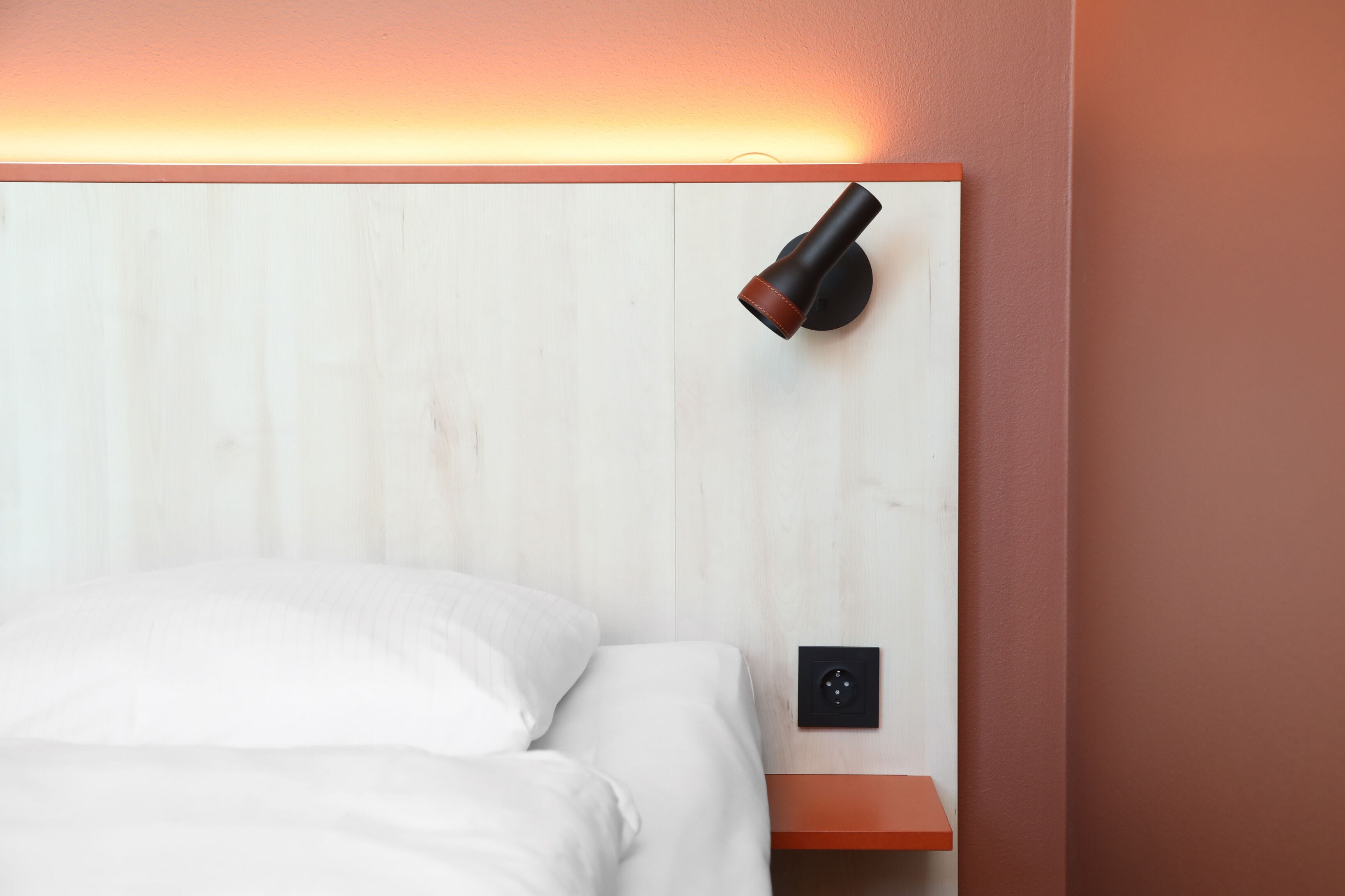 A neatly made bed with white linens, a wall-mounted nightstand with a black lamp, and a power outlet.