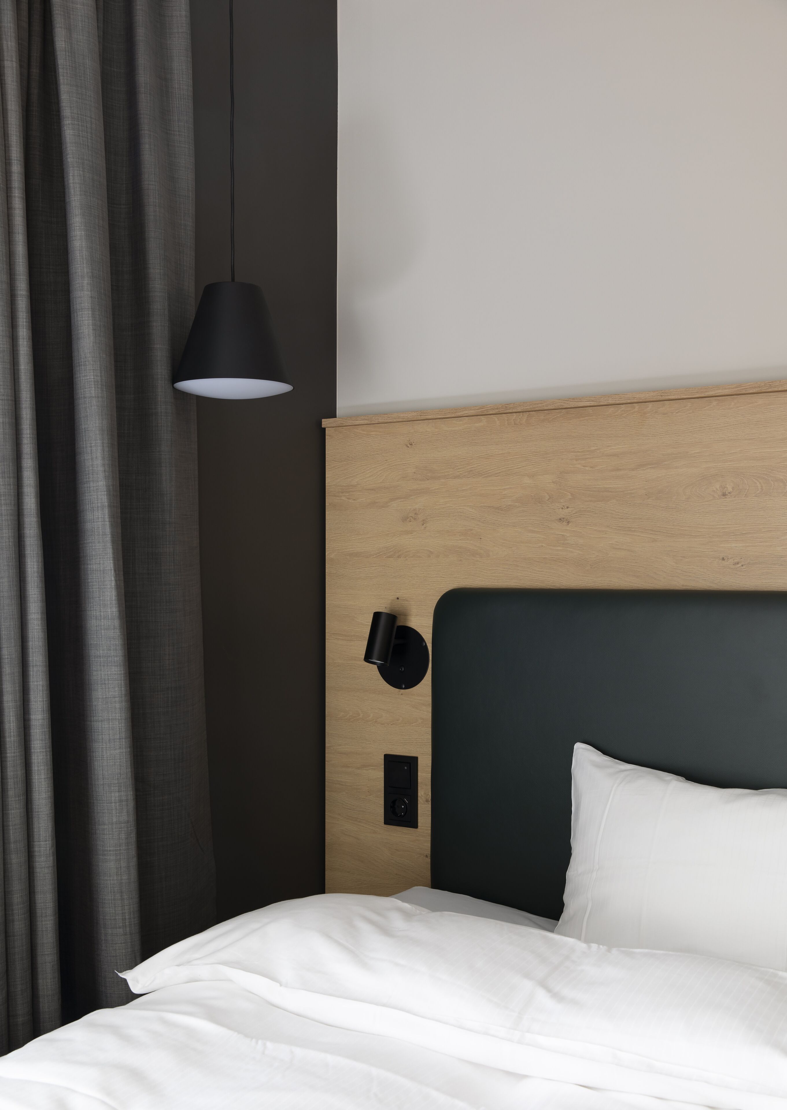 Una cama bien tendida con sábanas blancas, cabecero oscuro y una lámpara negra fijada a la pared.