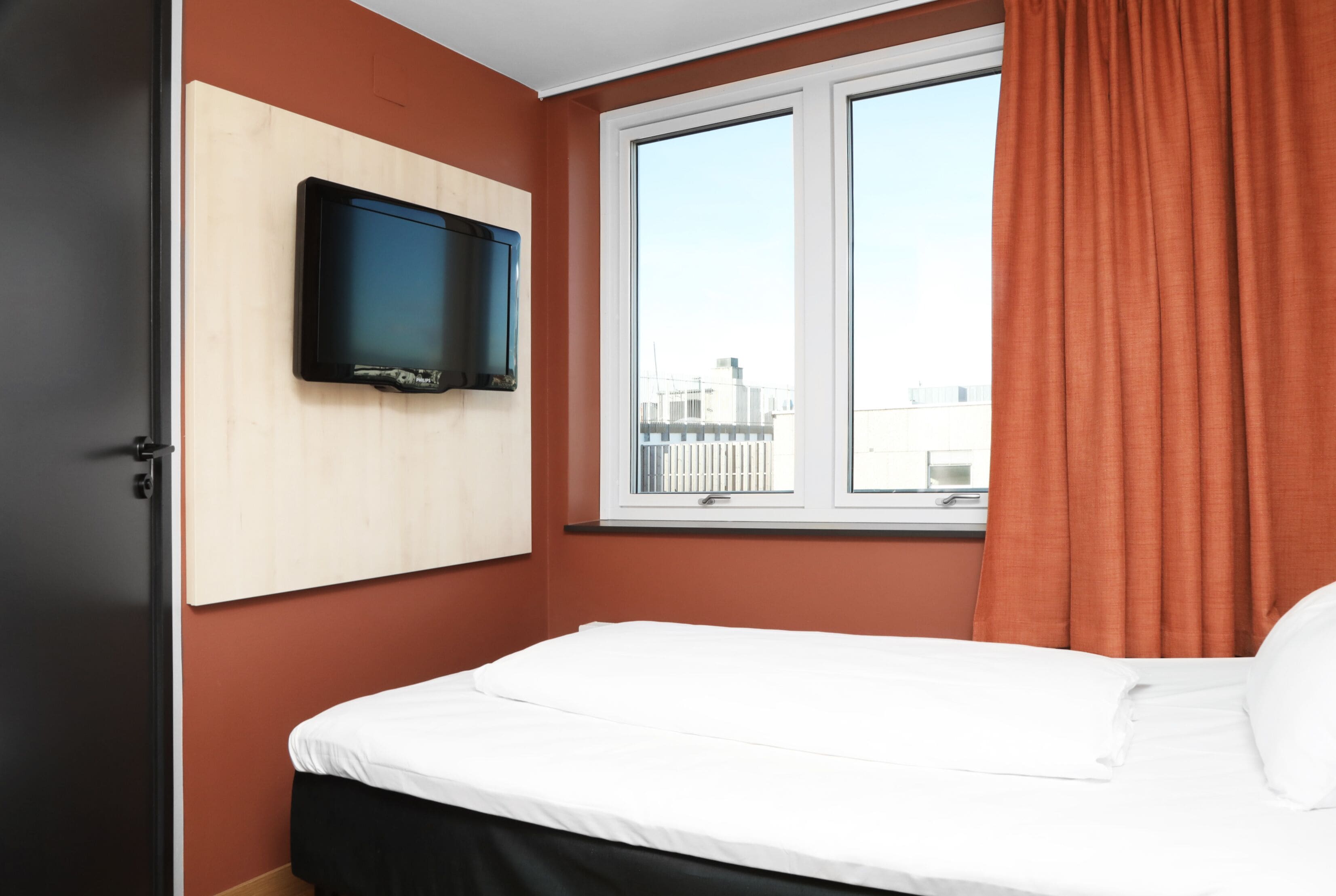 Ein Hotelzimmer mit Bett, einem an der Wand befestigten Fernseher, einem Fenster mit Vorhängen und Blick auf Gebäude.