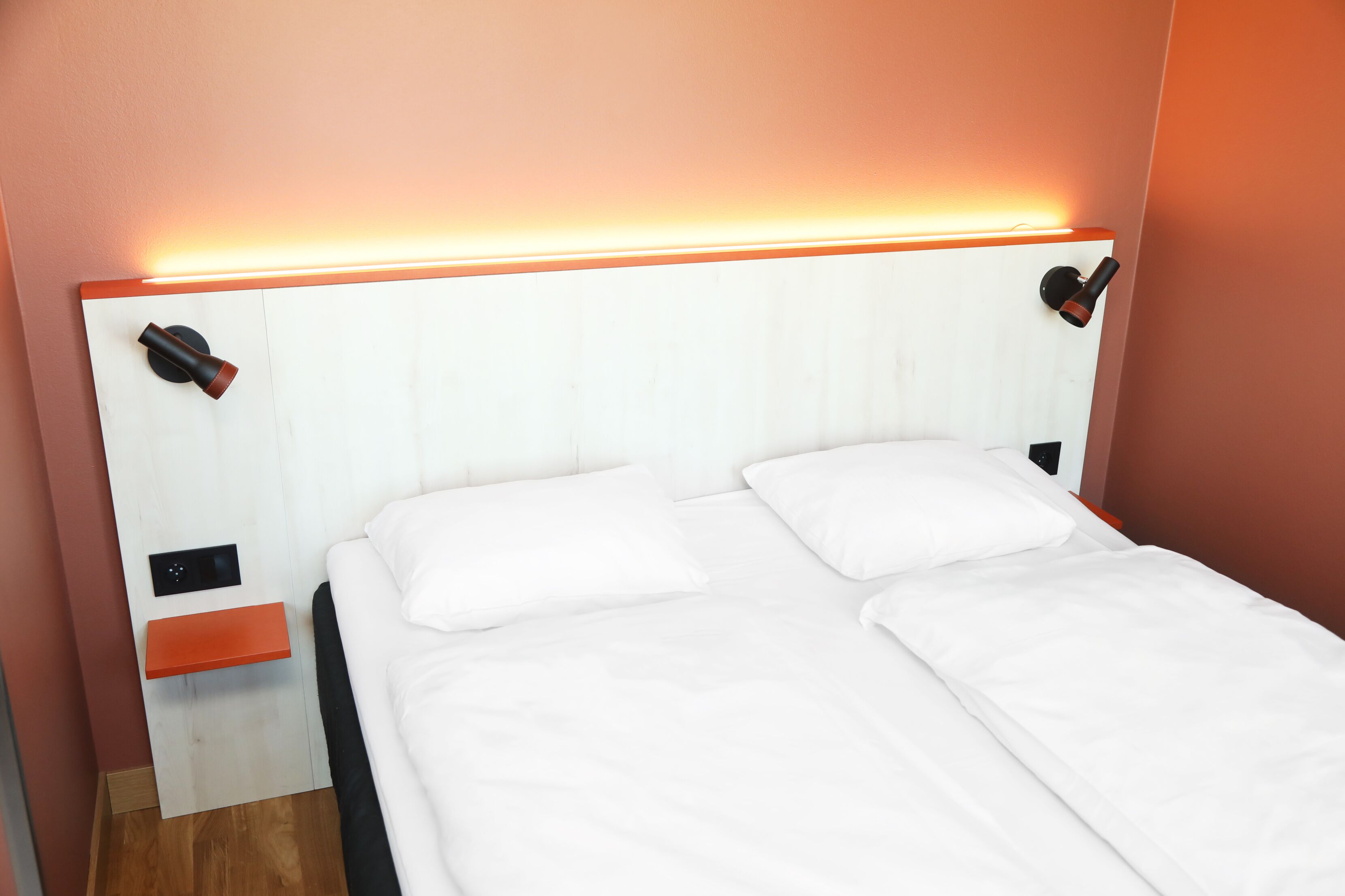 Un lit double avec du linge de lit blanc, une table de chevet en bois et un luminaire fixé au mur.