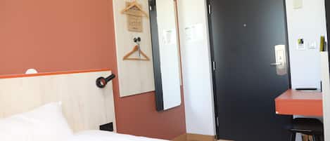 Habitación de hotel con cama, escritorio, silla, puerta y espejo adosado a la pared.