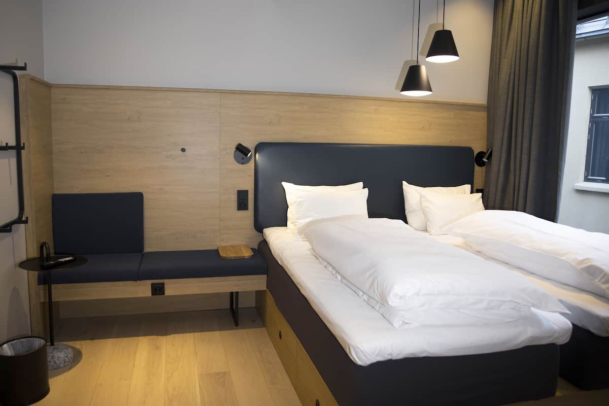 Une chambre d’hôtel moderne avec une tête de lit en bois, un lit avec du linge de maison blanc, une table de chevet, un banc et une lampe fixée au mur.