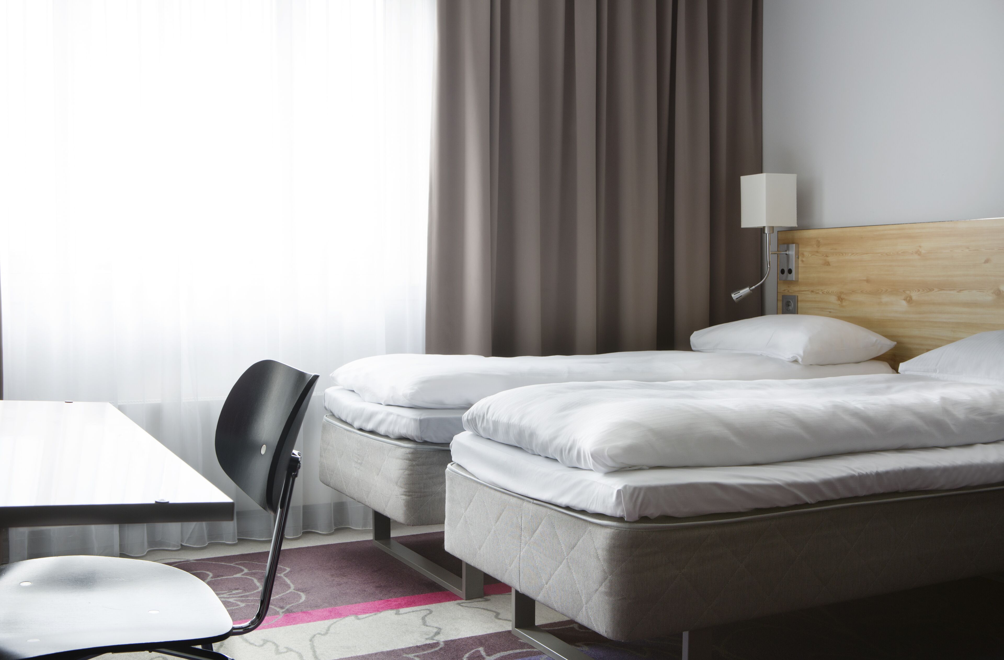 A hotel room with two single beds, a small table, a chair, and a lamp.