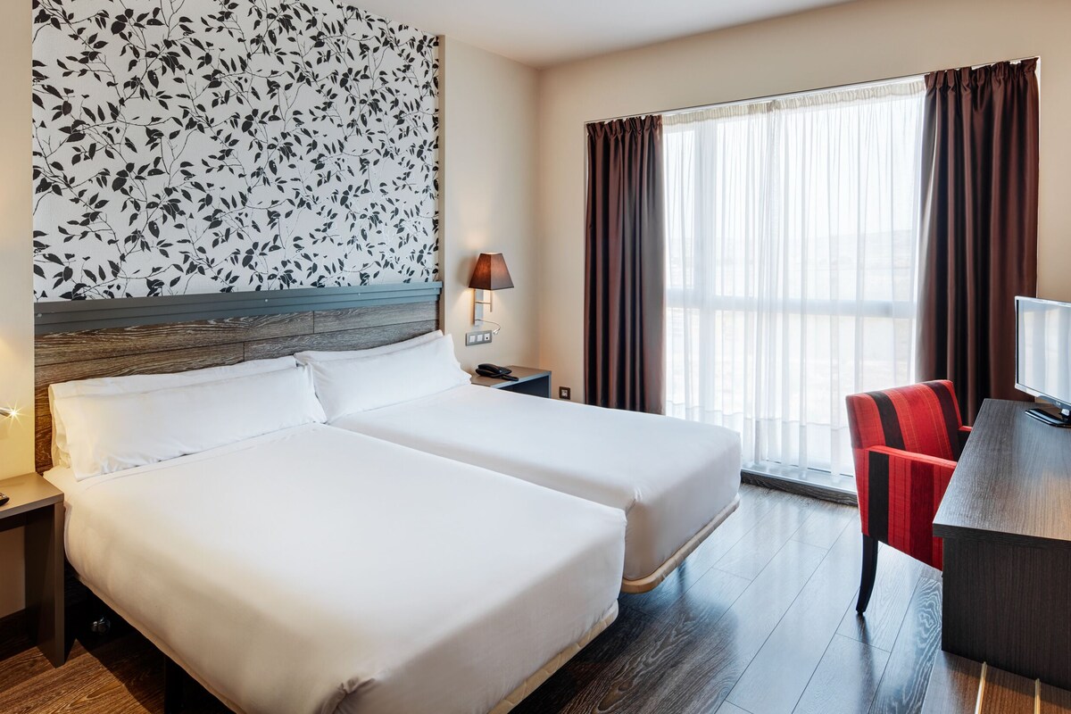 Une chambre d’hôtel avec deux lits, un bureau avec une télévision, une chaise et une fenêtre avec des rideaux.