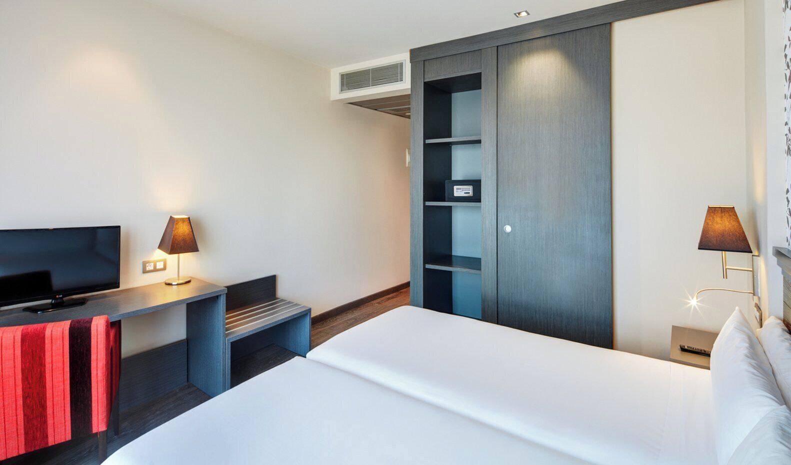 Una habitación de hotel moderna con una cama, un televisor, un escritorio con lámpara, un armario y una mesita de noche con lámpara.