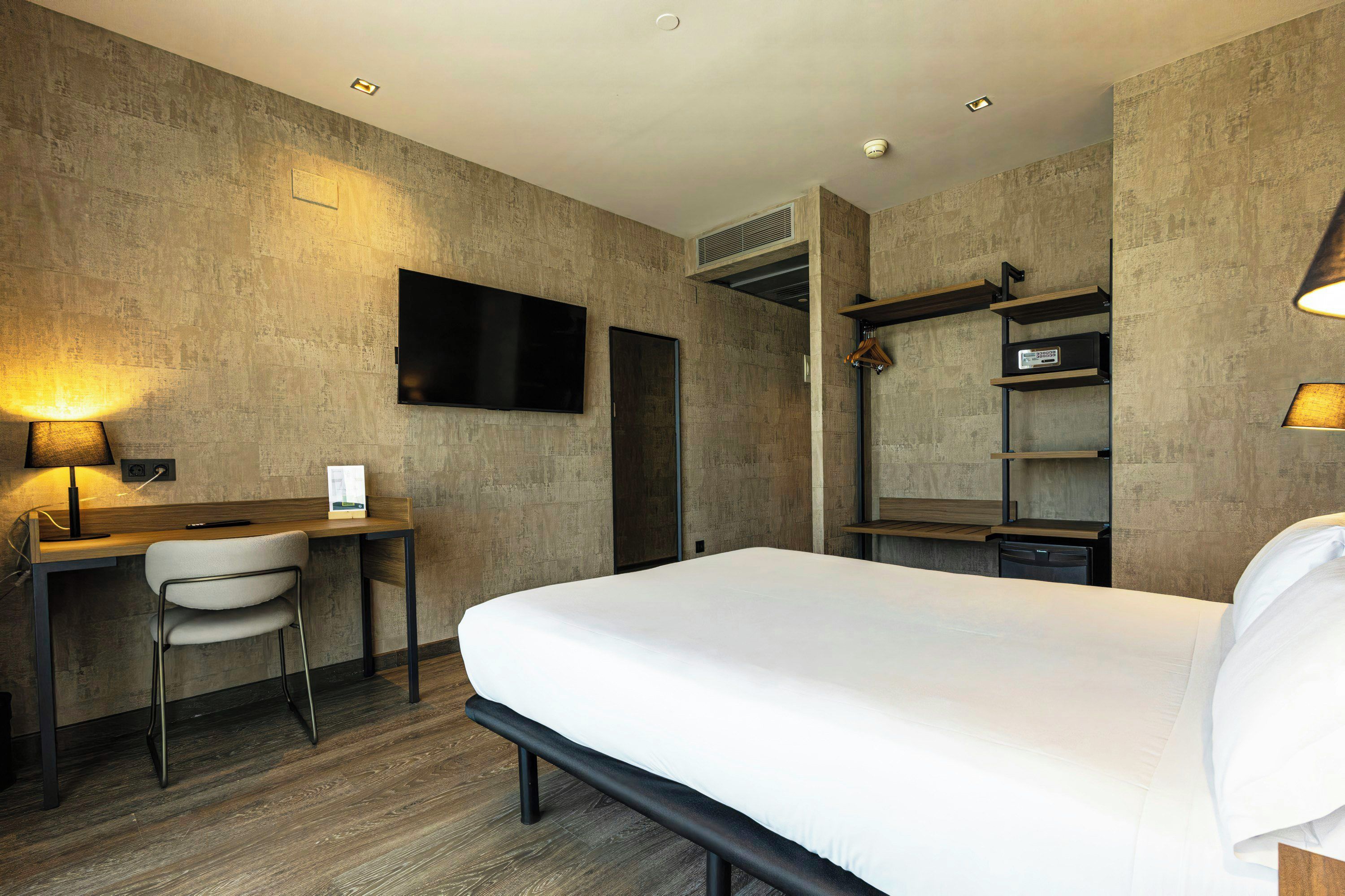 Ein Hotelzimmer mit Bett, Schreibtisch, Stuhl, Fernseher und einer Garderobe mit Regalen.