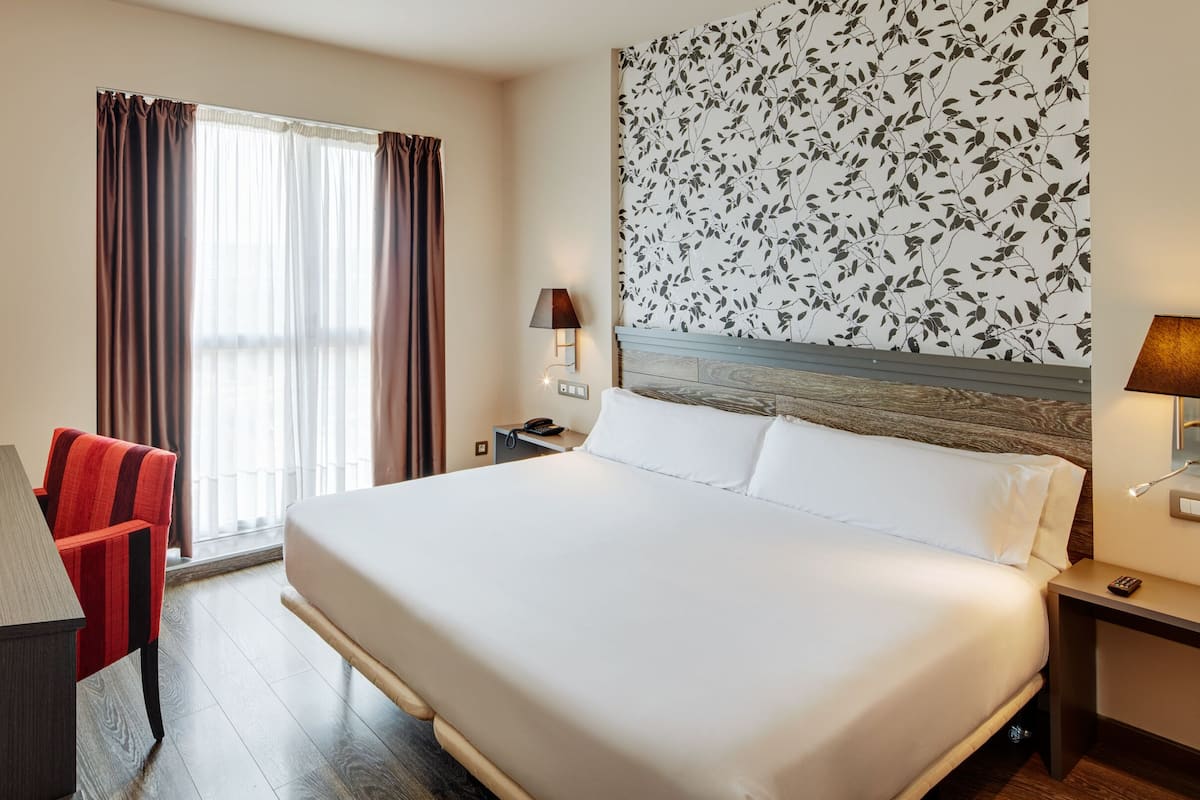 Une chambre d’hôtel avec un grand lit, un bureau, deux lampes, une chaise et une fenêtre avec des rideaux.