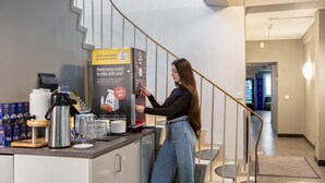A woman at a coffee station with a coffee machine, a coffee pod dispenser, and a sign advertising coffee specialties and hot drinks. - a&o Köln Hauptbahnhof (Cologne)