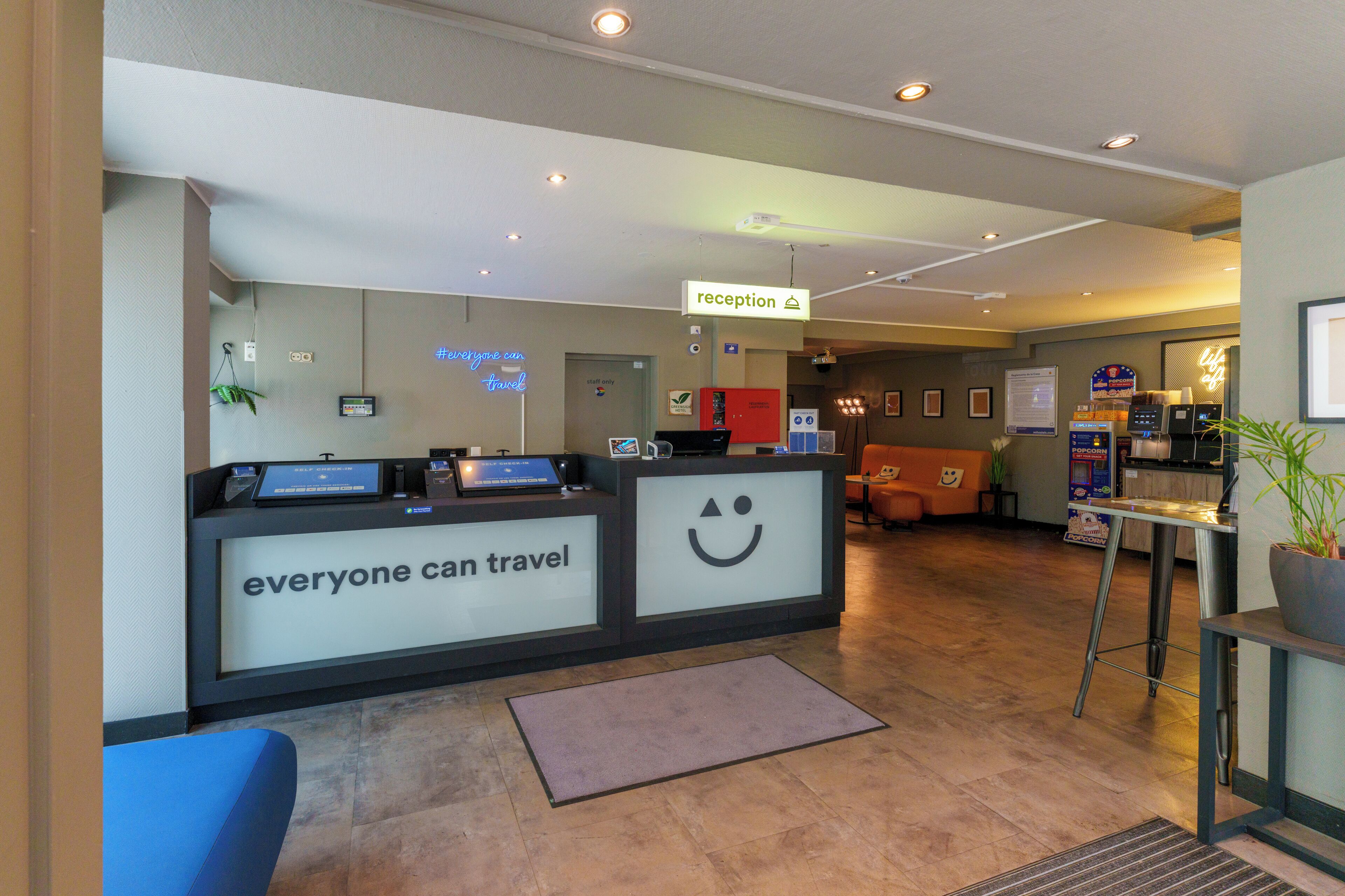 Un'area reception con una stazione di check-in automatico, una scrivania della reception con una faccina sorridente e un'area relax con una macchina per popcorn.