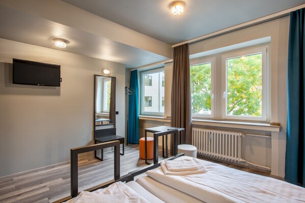 A modern hotel room with a bed, a desk, a chair, a mirror, a TV, and a window with curtains.