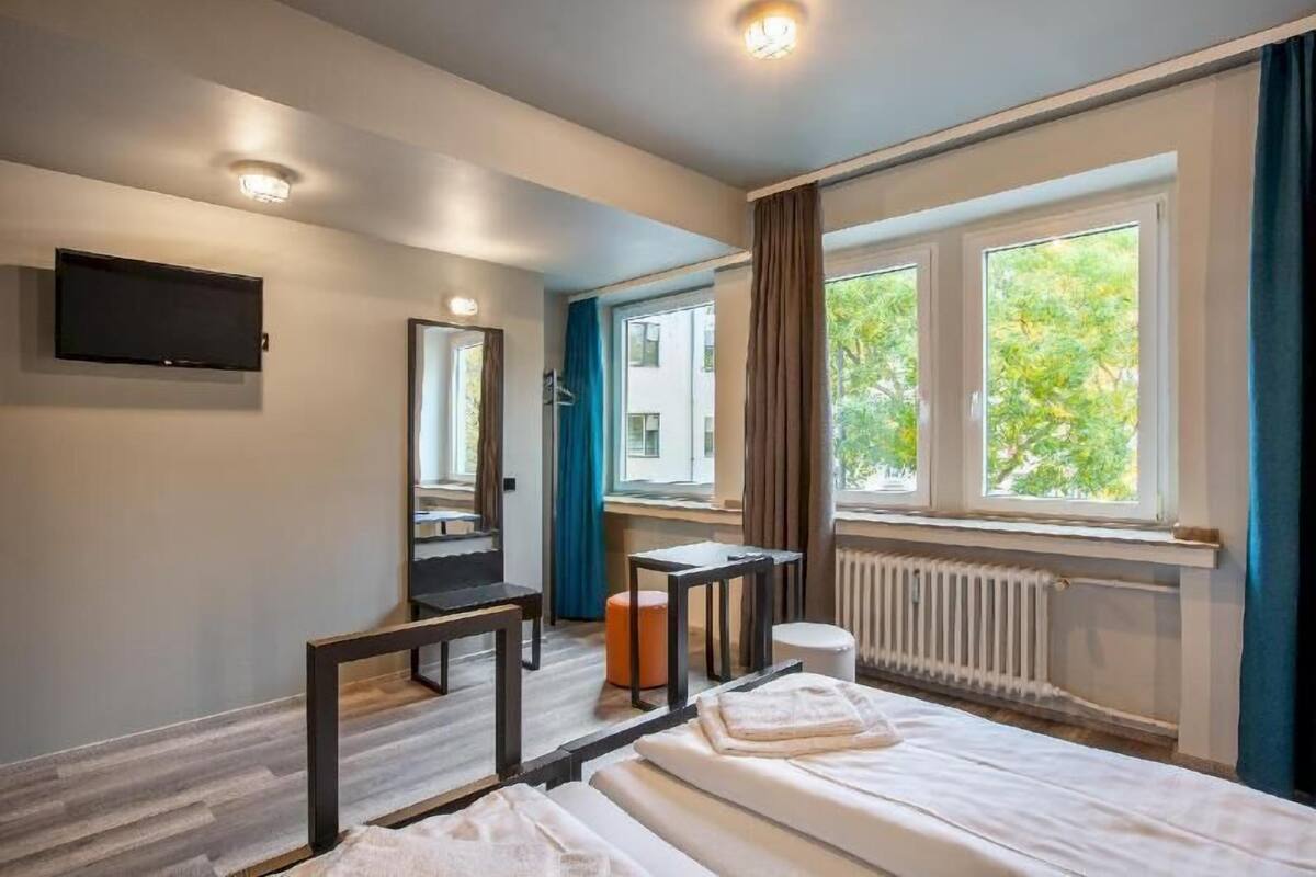 Habitación de hotel con cama, escritorio, espejo, televisor y ventana con vista a árboles.