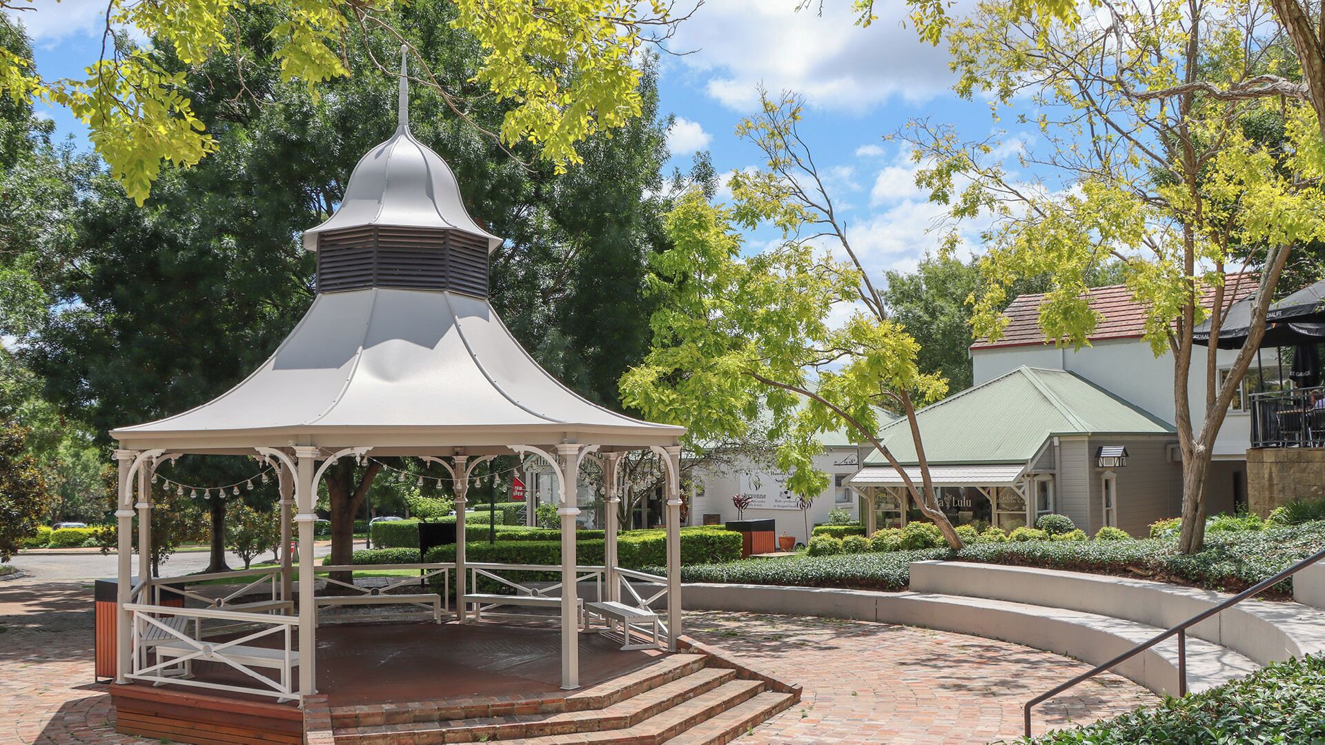 Un padiglione con tetto a cupola e piattaforma in legno, immerso in un giardino curato con alberi e un edificio sullo sfondo.