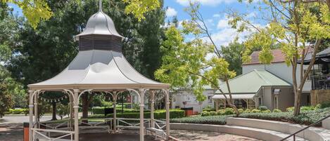Un padiglione con tetto a cupola e piattaforma in legno, immerso in un giardino curato con alberi e un edificio sullo sfondo.