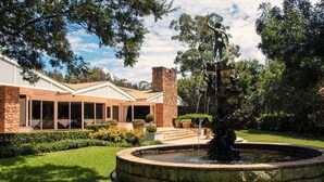 Un patio con una fuente, un edificio de piedra con amplios ventanales y un césped bien cuidado.