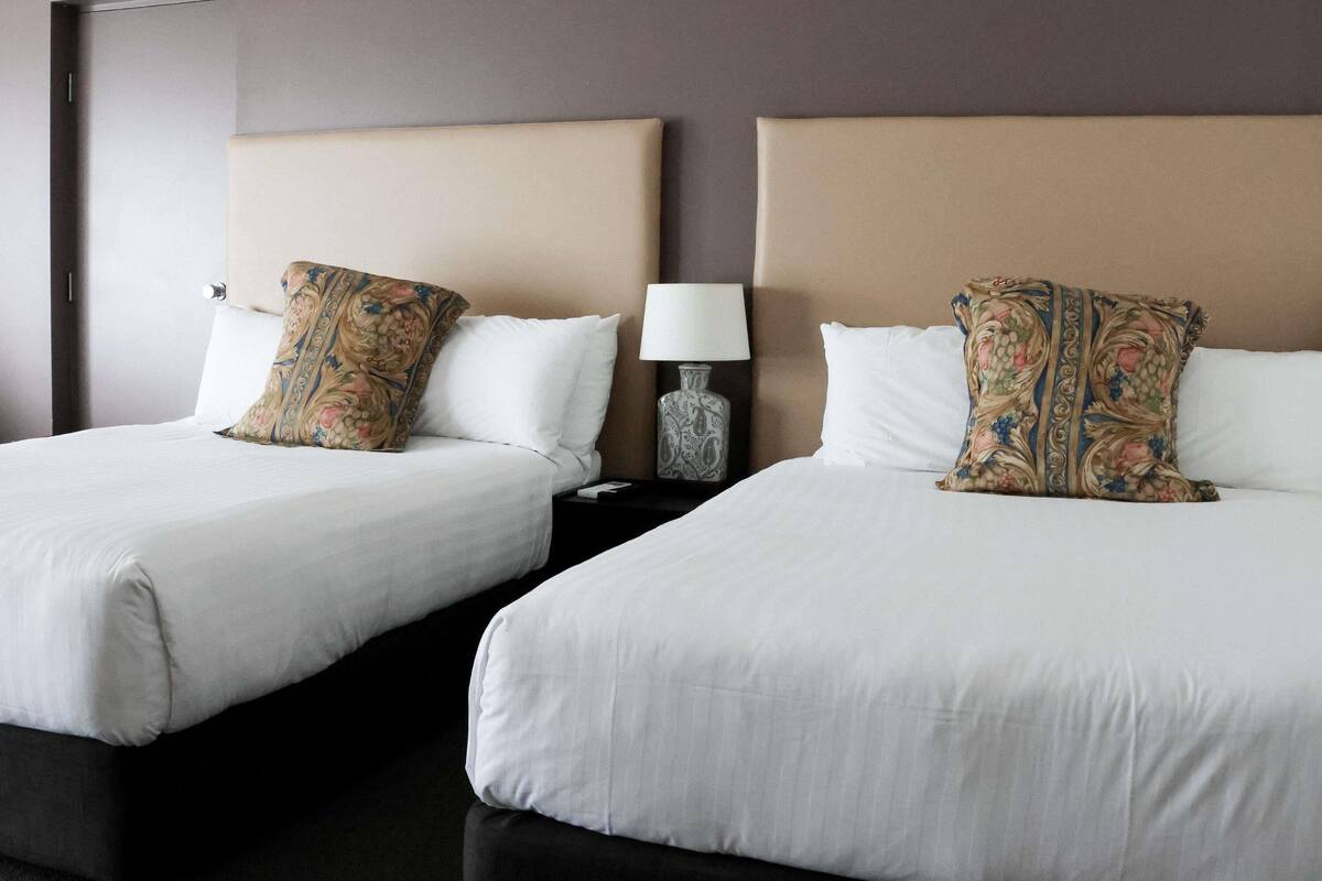 Two beds with white linens, a beige headboard, and a nightstand with a lamp and decorative vase.