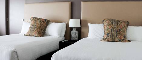 Two beds with white linens, a beige headboard, and a nightstand with a lamp and decorative vase.