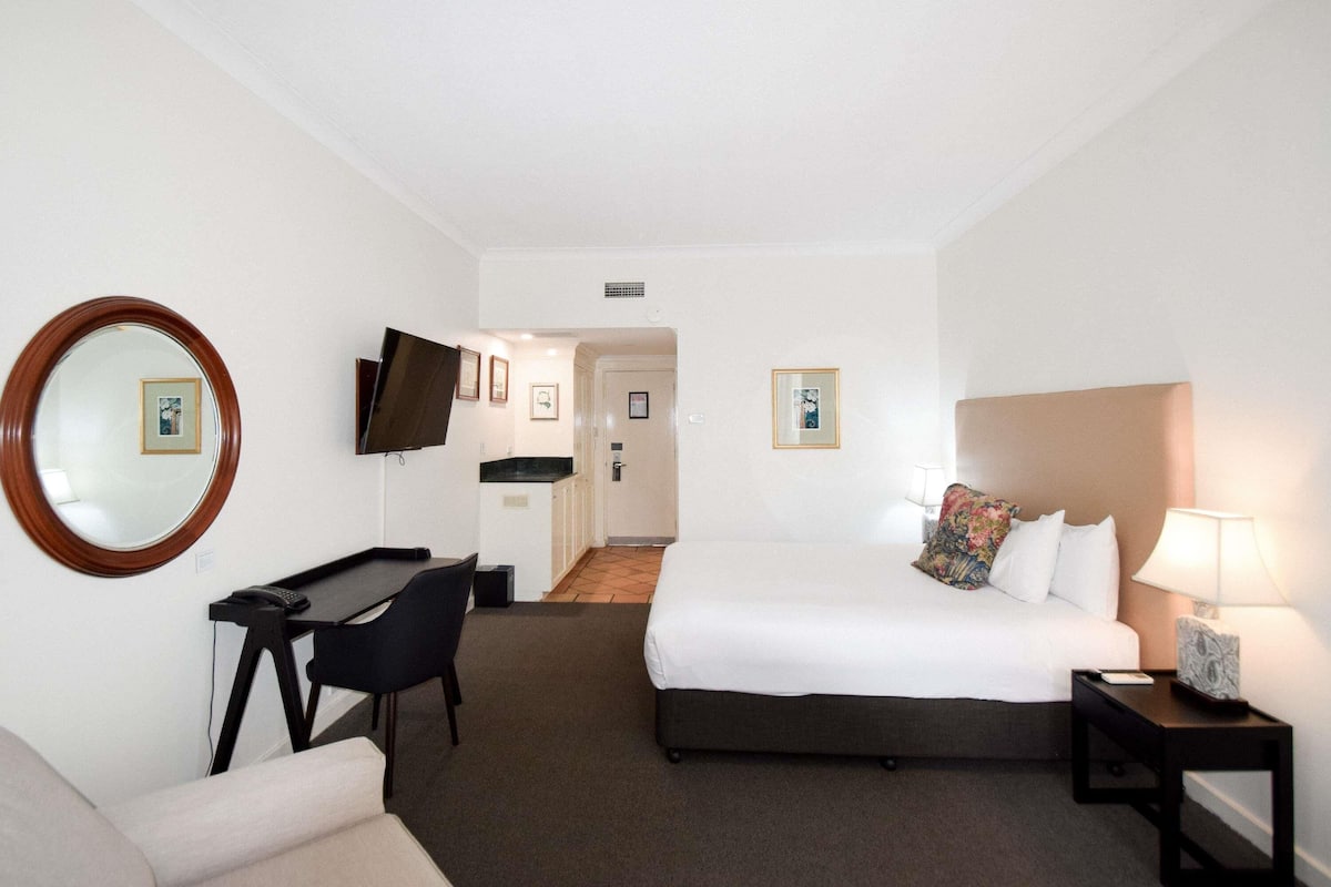 A hotel room with a bed, a desk, a chair, a TV, a bedside table with a lamp, and a mirror.