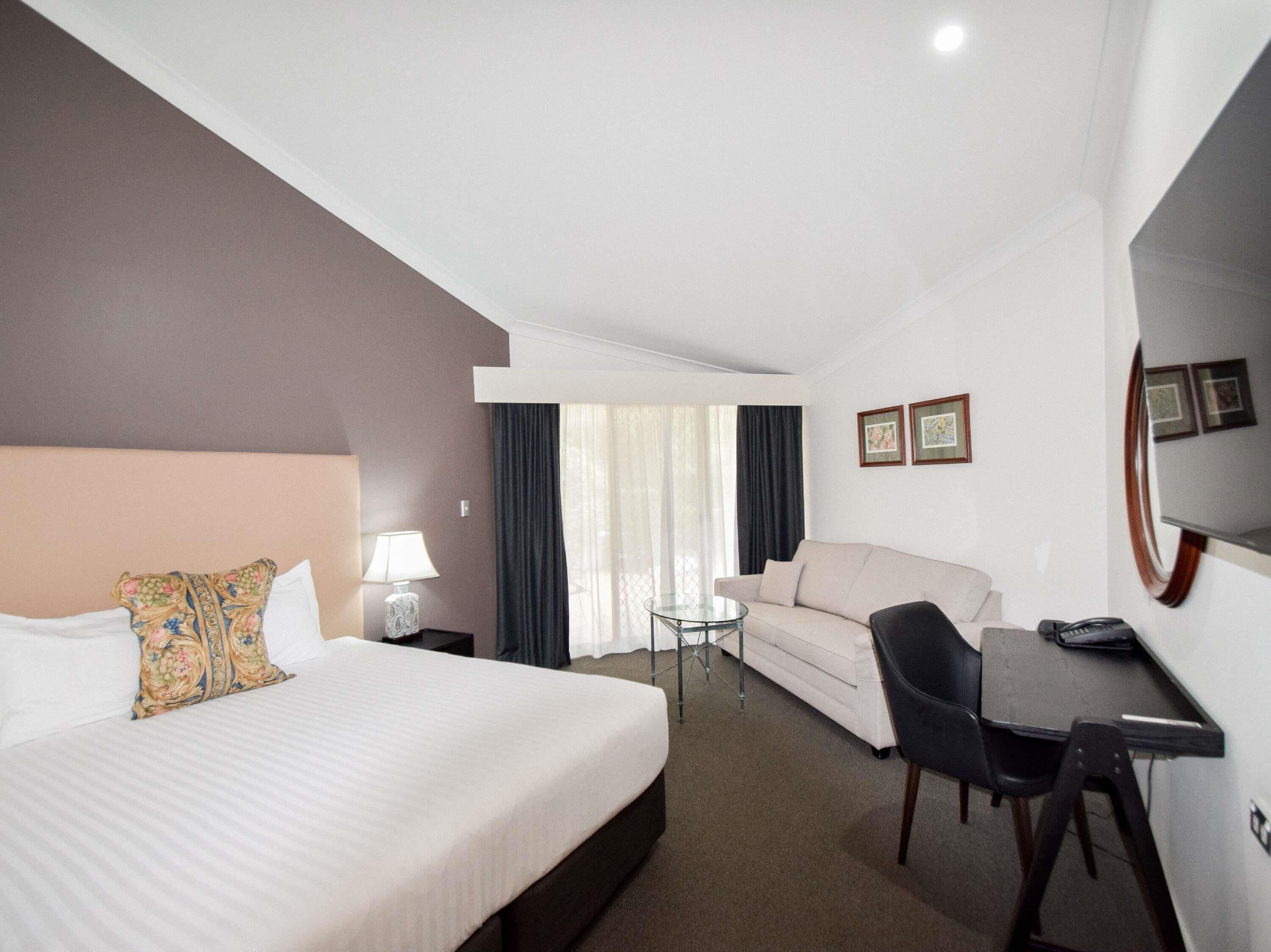 Una habitación de hotel con cama, sofá, escritorio, silla, lámpara, mesa de vidrio y ventana con cortinas.