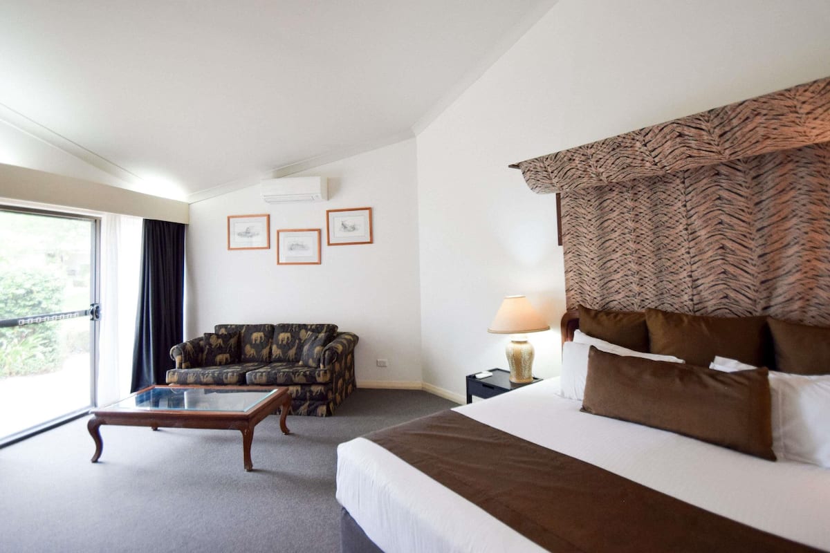 A hotel room with a bed, a sofa, a glass coffee table, a lamp, and three framed pictures on the wall.