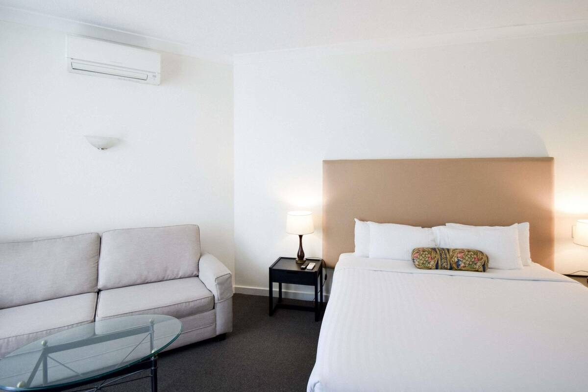 A hotel room with a bed, a sofa, a glass coffee table, a nightstand with a lamp, and an air conditioning unit on the wall.