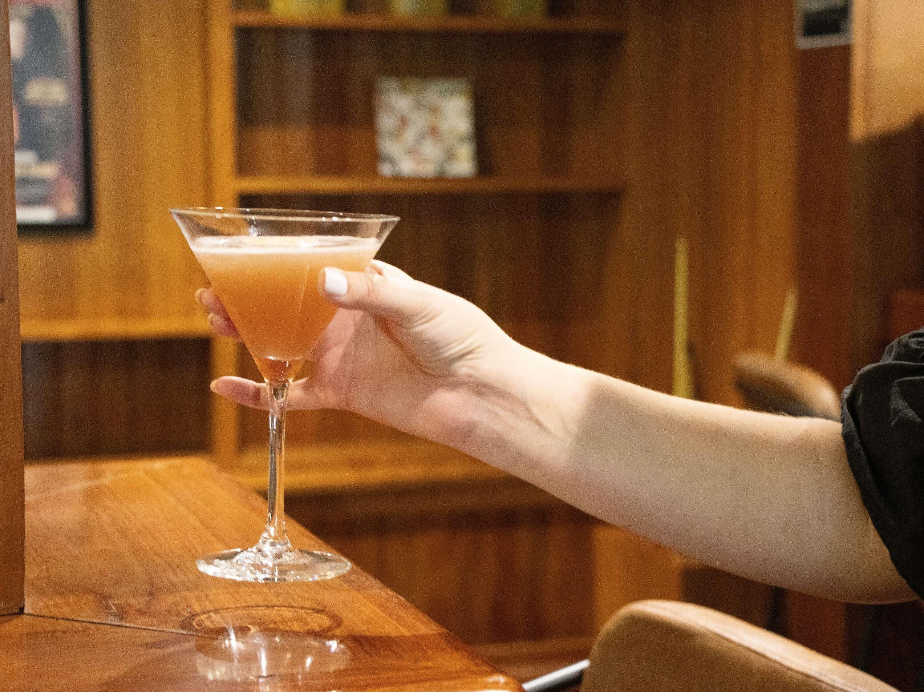 Une main tenant un verre à martini rempli d’un liquide transparent, le tout sur un fond en bois avec des étagères.