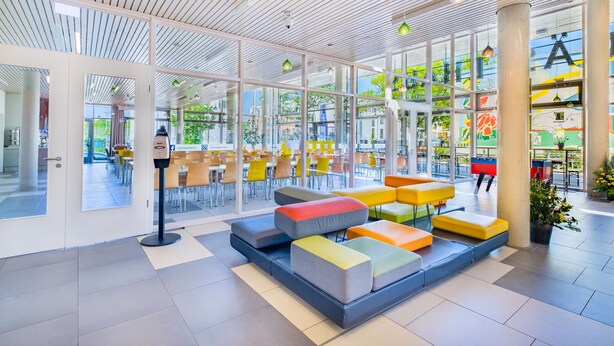 A spacious indoor area with a modern design, featuring a seating area with colorful cushions, large windows, and a view of the outside.