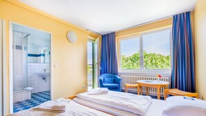 A hotel room with two beds, a bathroom, a small table, a chair, and a view of greenery outside.