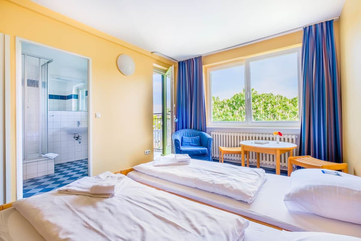 A hotel room with two beds, a bathroom, a small table, a chair, and a view of greenery outside.