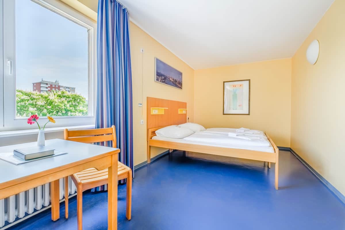 A hotel room with a bed, a desk with a chair, a window with curtains, and a radiator.