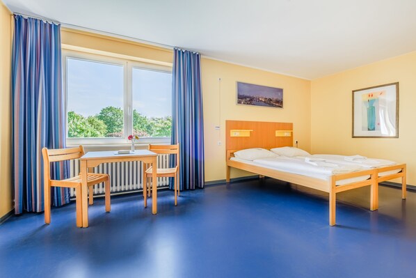 Habitación de hotel con una cama, un escritorio y dos sillas, una ventana con cortinas, un radiador y un cuadro de un paisaje urbano en la pared.