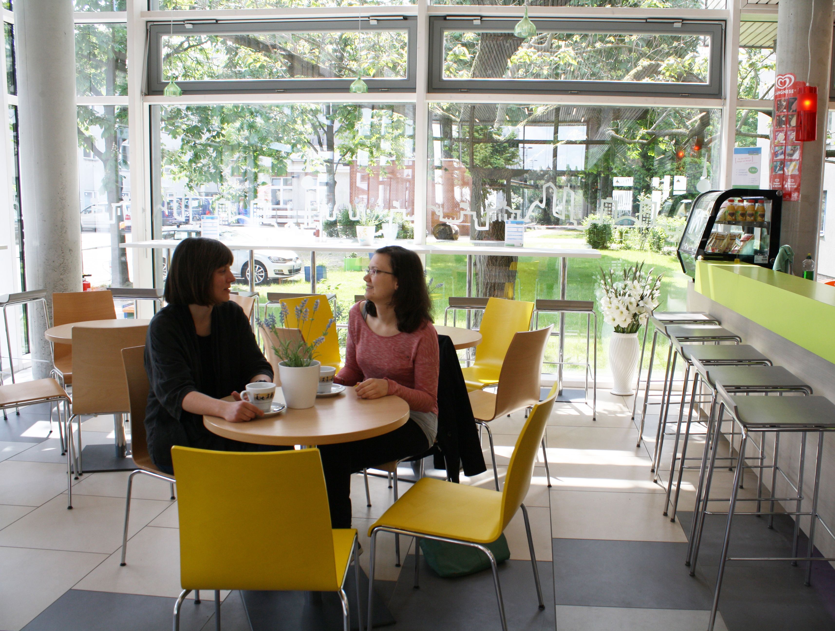 Una cafetería con dos mesas, sillas amarillas y grandes ventanales que dan a la calle y a zonas verdes.