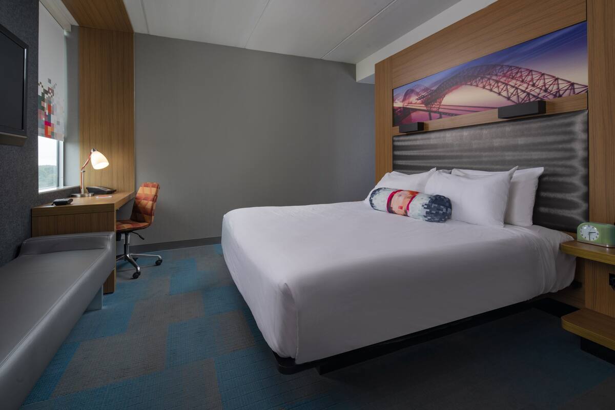 A hotel room with a large bed, a desk with a lamp, a chair, and a painting of a bridge on the wall.