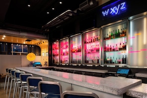 A bar area with a neon sign, a digital display, and a row of chairs. - Aloft by Marriott Rogers-Bentonville (Rogers)