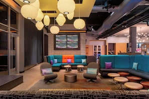 A modern lounge area with a sectional sofa, armchairs, and ottomans, featuring a patterned rug and pendant lighting. - Aloft by Marriott Rogers-Bentonville (Rogers)