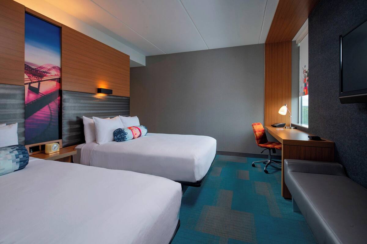 A hotel room with two beds, a desk, a chair, and a TV.