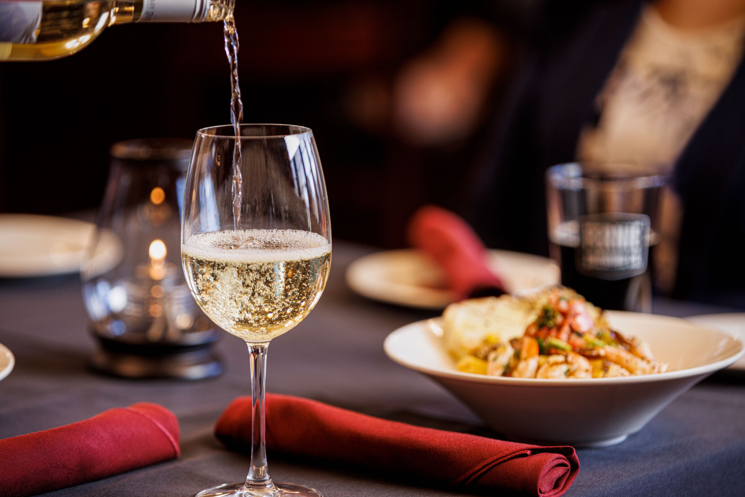 Un verre de vin blanc est versé dans un verre à pied, un bol de pâtes aux légumes et une serviette rouge sont posés sur une table.