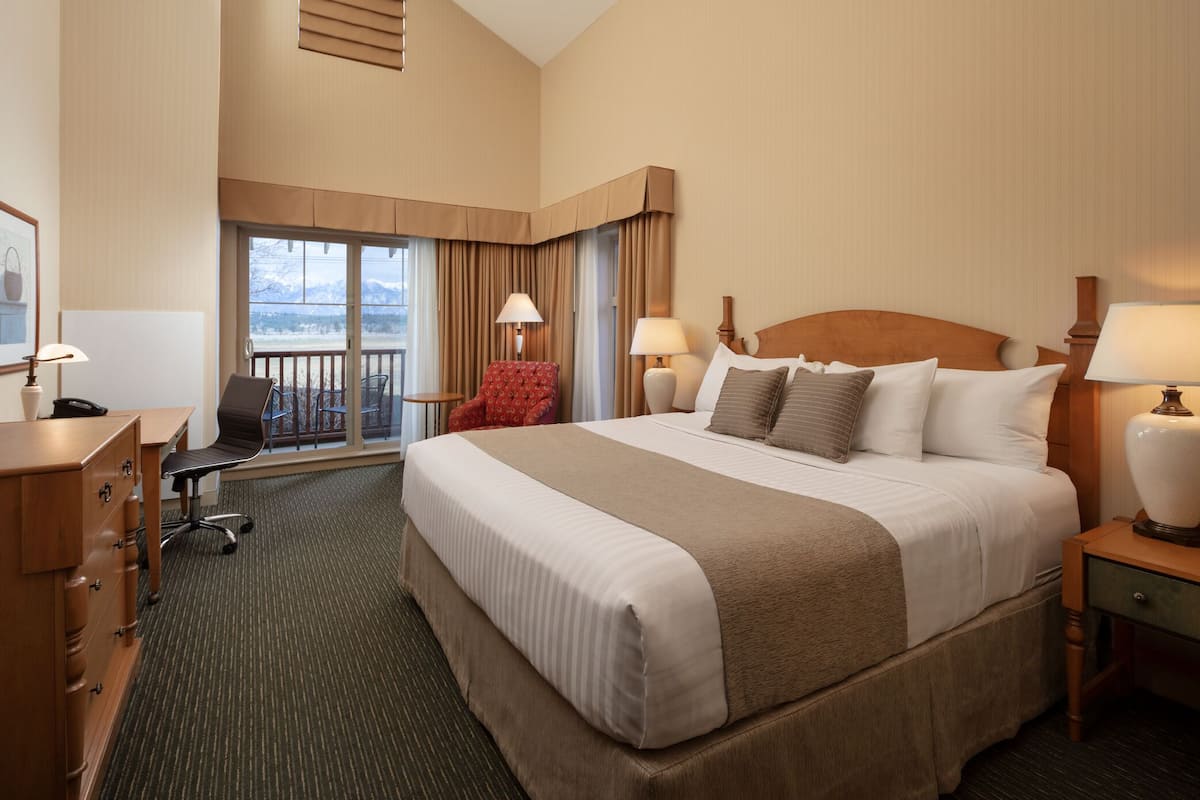 A hotel room with a large bed, a desk with a chair, a red armchair, and a balcony with a view of mountains.