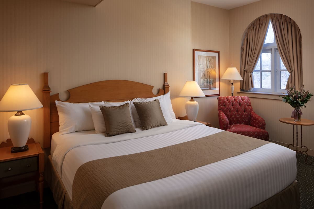A neatly made bed with a wooden headboard, two bedside lamps, a red armchair, and a small table with a vase of flowers.