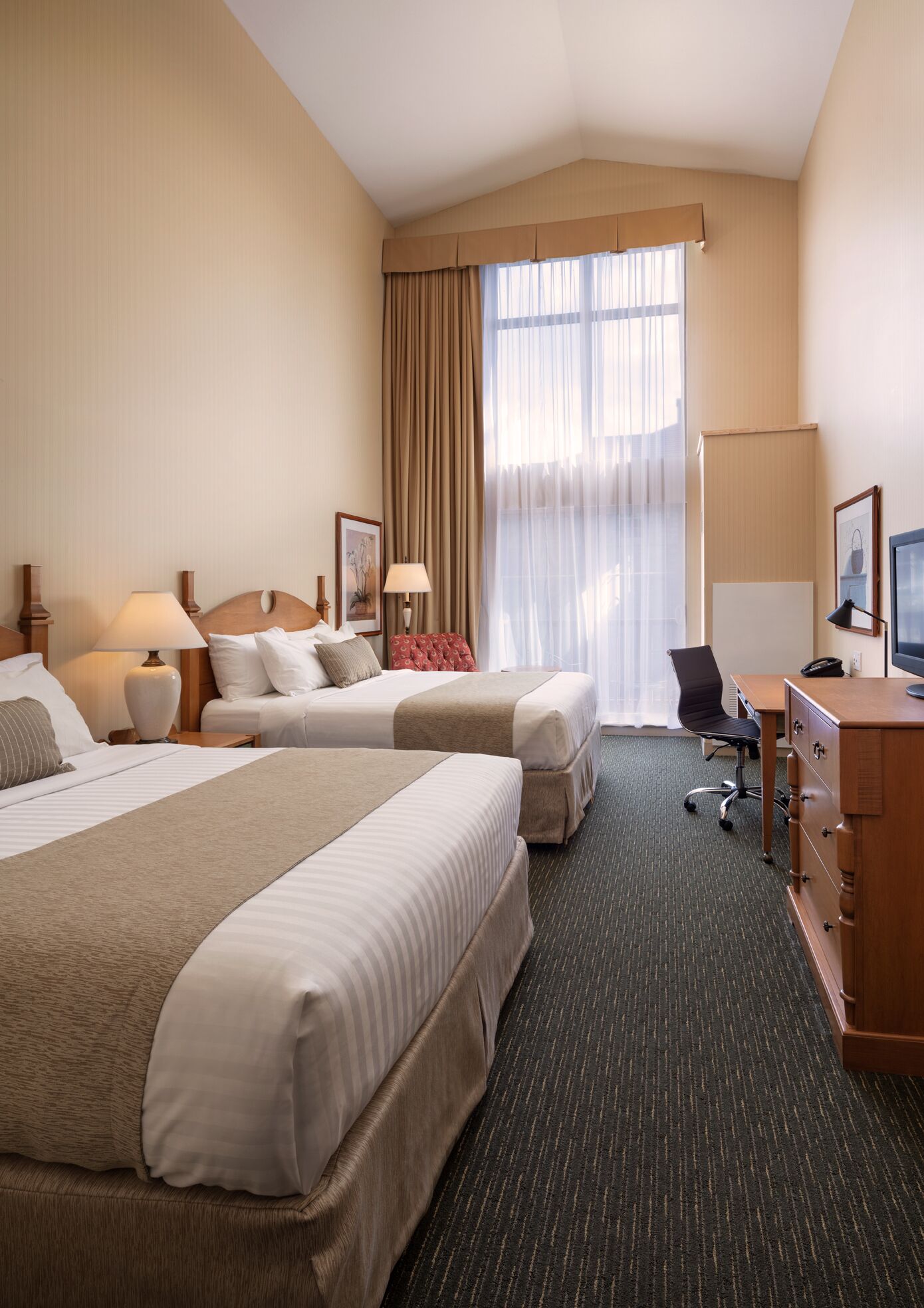 Quarto de hotel com duas camas, uma escrivaninha, uma cadeira, uma televisão e uma janela com cortinas.