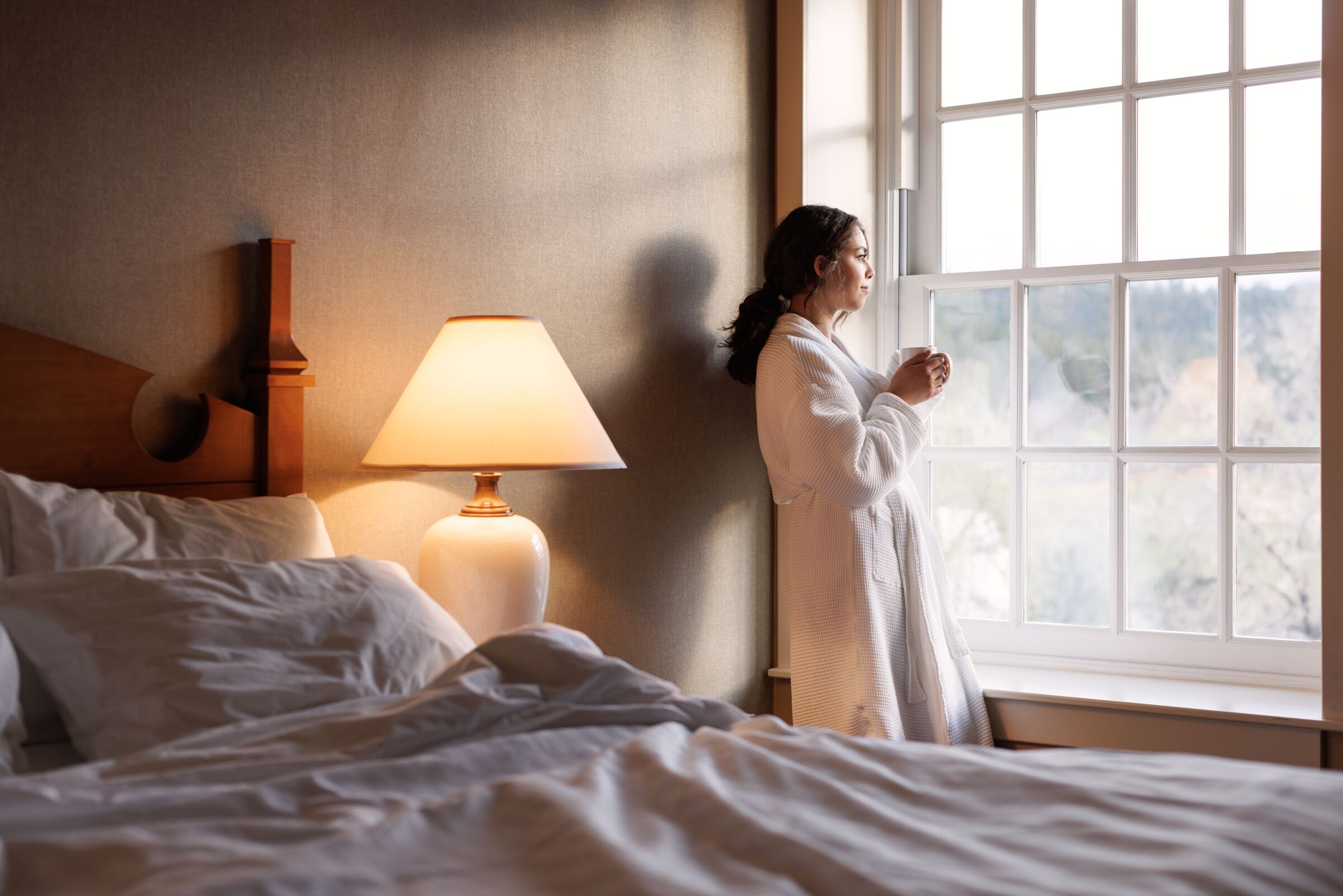 Habitación de hotel con una cama, una mesita de noche con lámpara y una ventana con vistas a árboles.