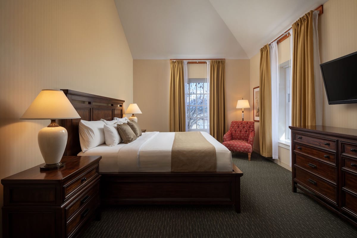 A hotel room with a large bed, two bedside tables with lamps, a red armchair, a TV, and a window with curtains.