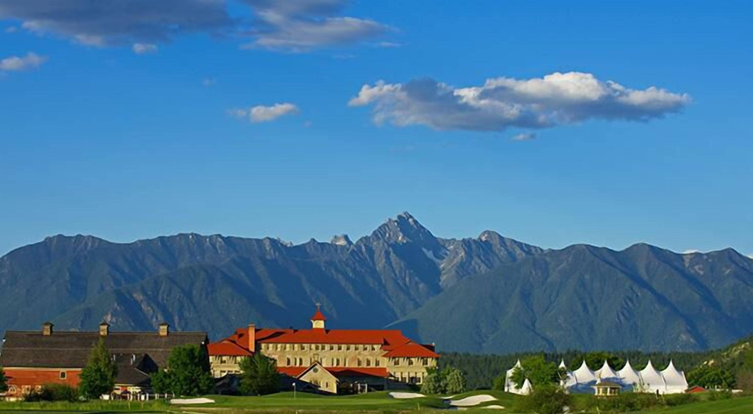 Un grand bâtiment à toit rouge, entouré d’une pelouse bien entretenue et de tentes, avec une chaîne de montagnes en arrière-plan.