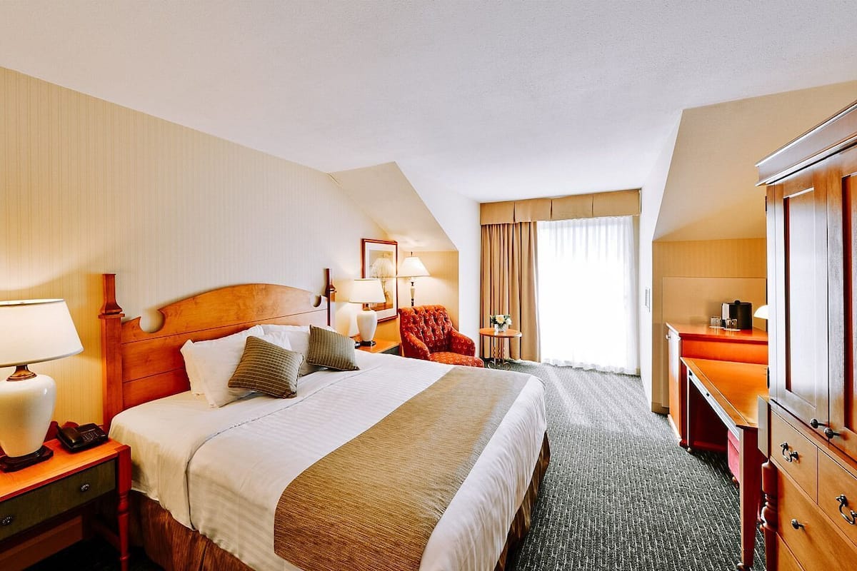 A hotel room with a large bed, a desk with a lamp, a chair, a dresser, and a window with curtains.