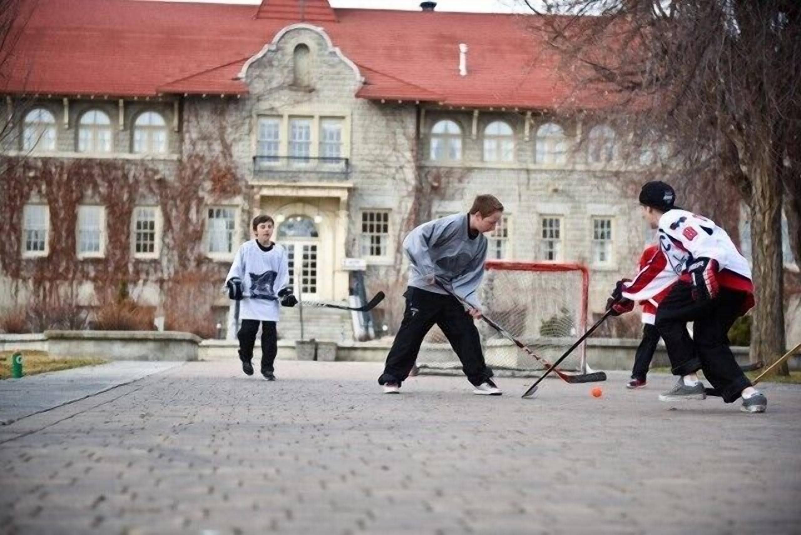 A group of people playing hockey on an outdoor court with a stone building in the background.