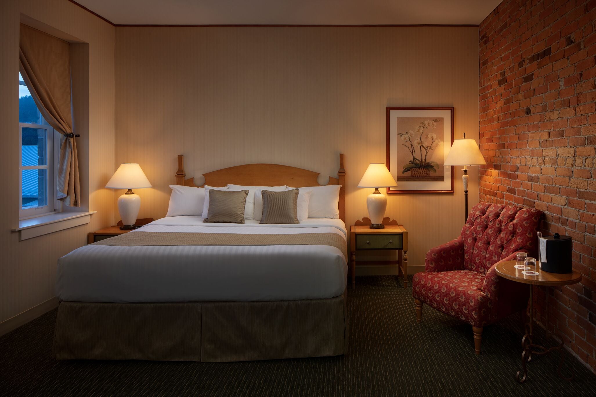 Une chambre d’hôtel avec un grand lit, deux tables de chevet avec des lampes, un fauteuil rouge et un mur en briques.